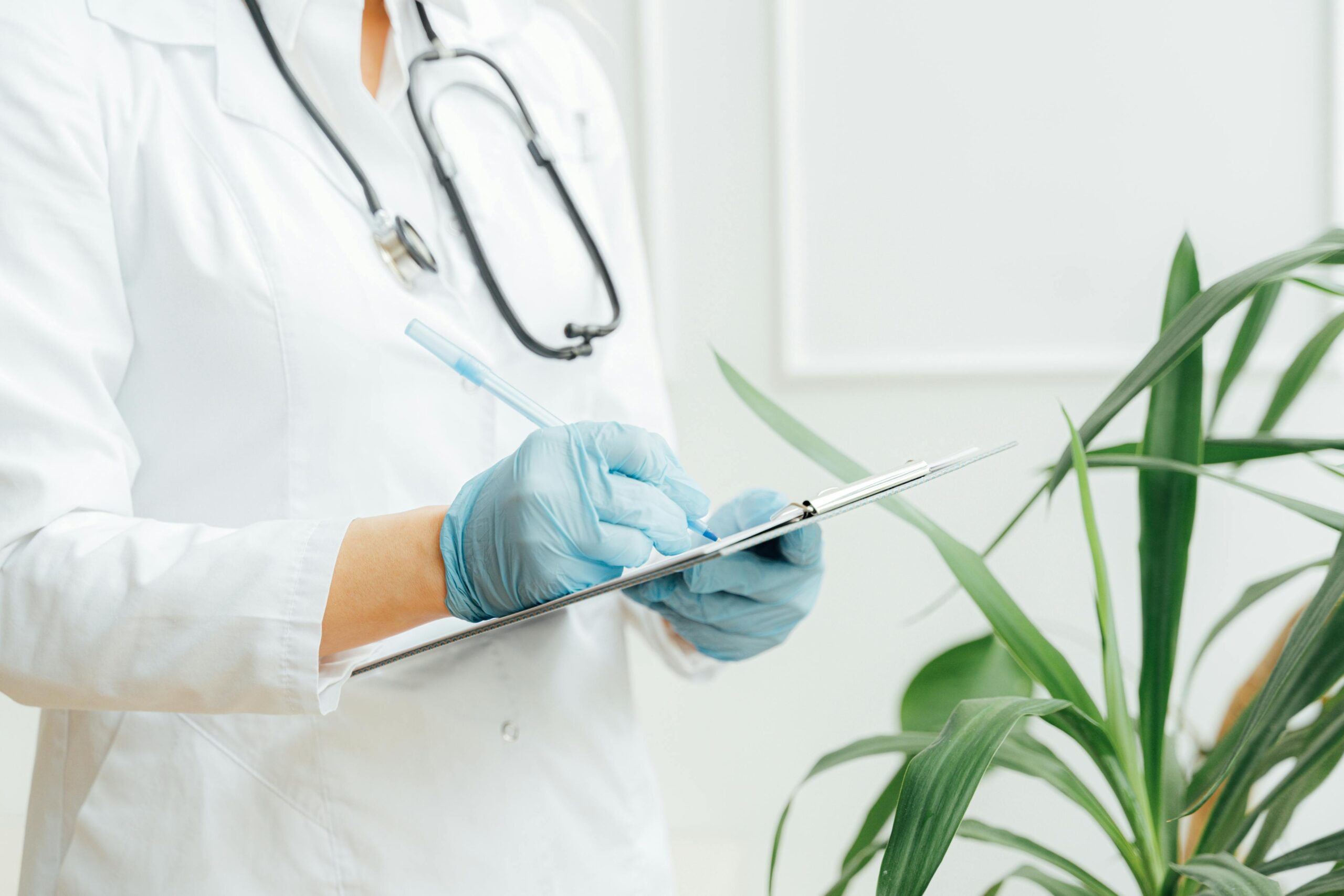 A healthcare professional wearing gloves takes notes on a clipboard with a stethoscope around their neck.