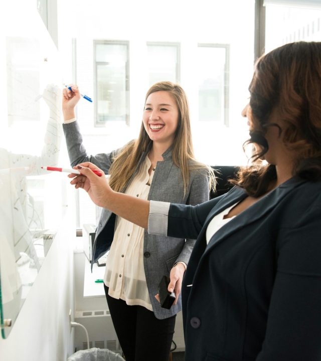 Two professional women discussing ideas on a whiteboard in a modern office setting.